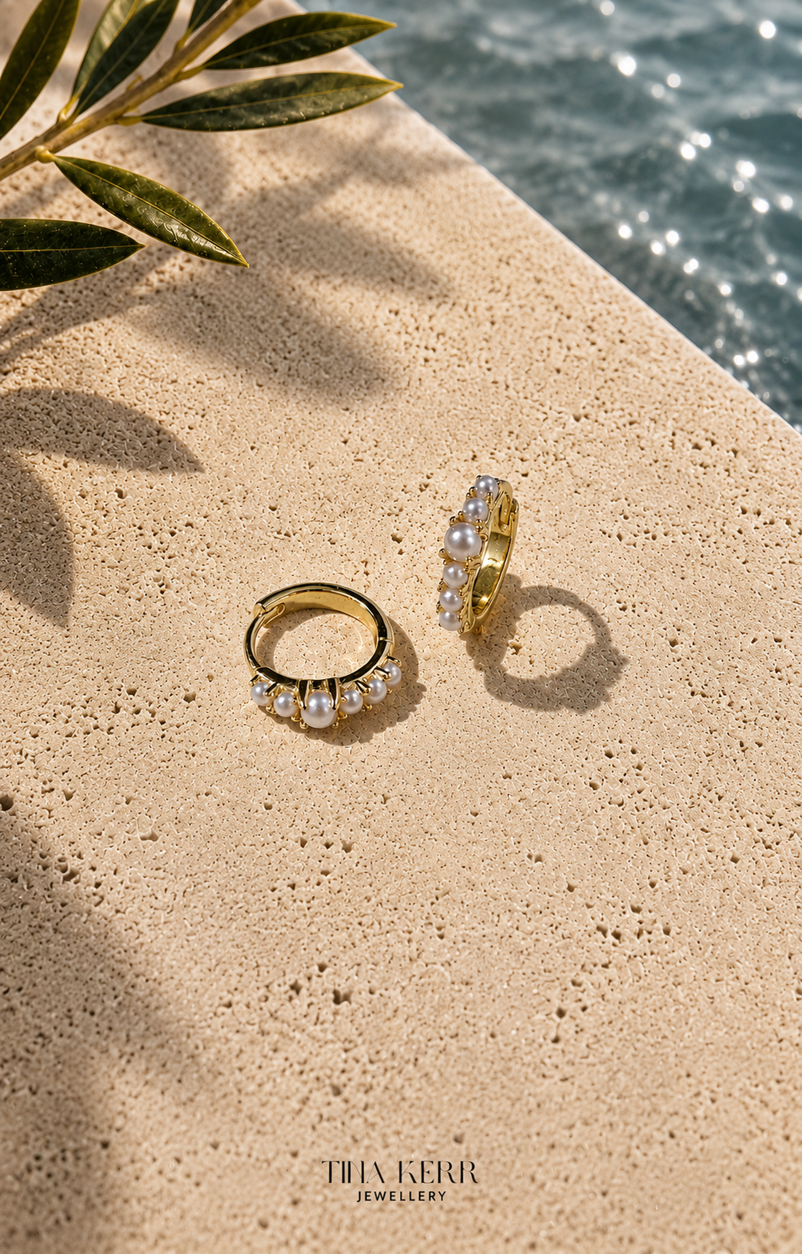 Gold and pearl earrings on a sandy surface with water and leaves in the background, THK Kerr Jewellery branding visible.