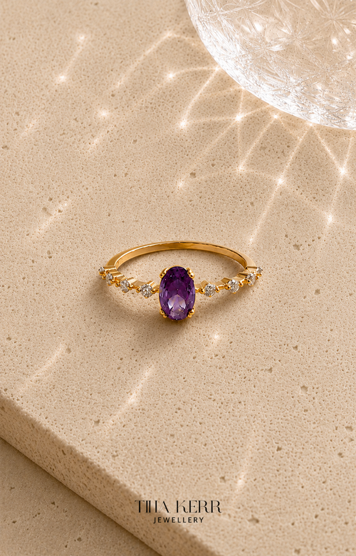 Gold ring with an amethyst stone on a beige surface, branded 'Tina Kerr Jewellery'.