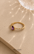Gold ring with a purple gemstone on a beige surface, featuring the brand 'Tina Kerr Jewellery'.