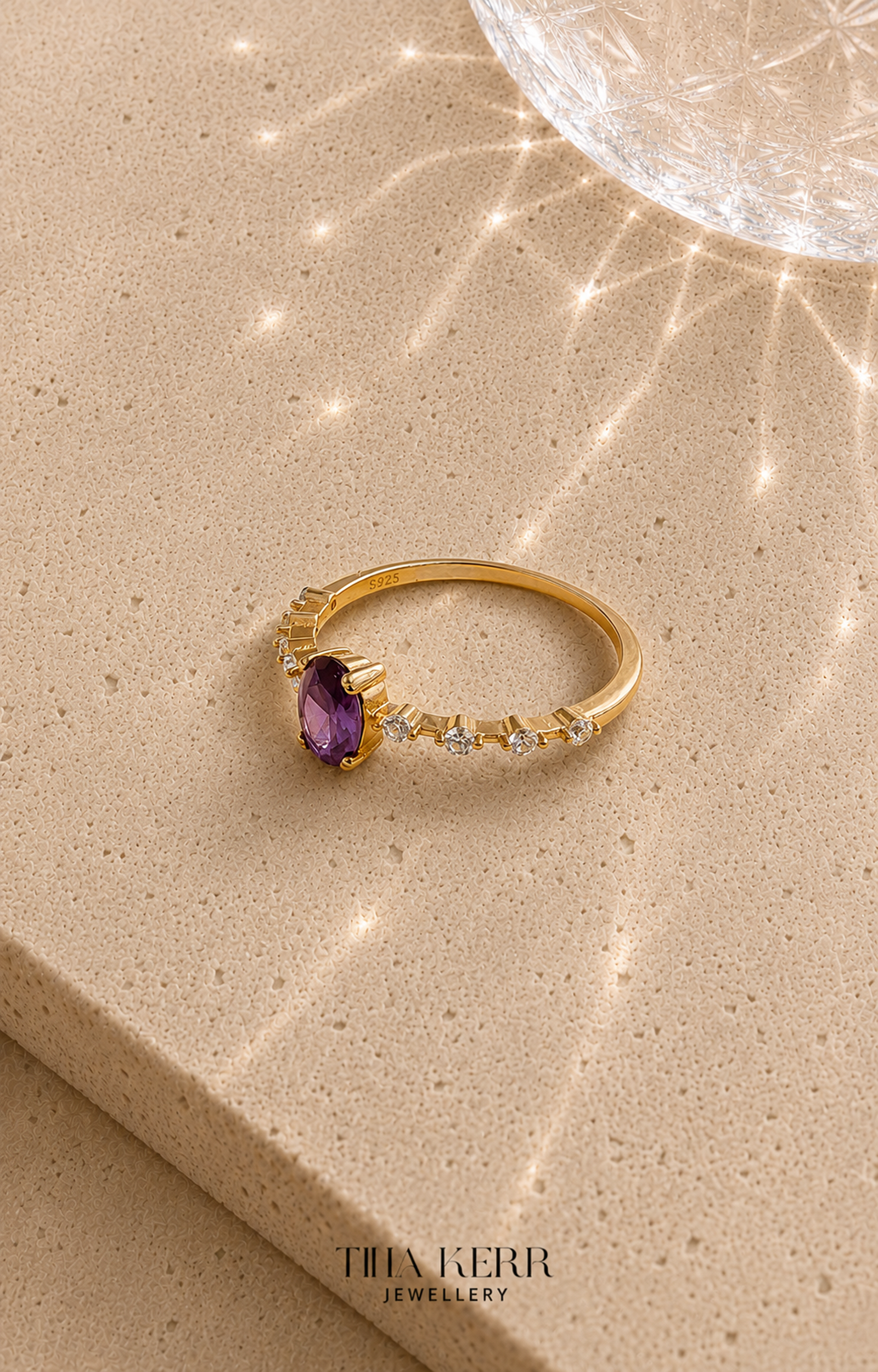 Gold ring with a purple gemstone on a beige surface, featuring the brand 'Tina Kerr Jewellery'.